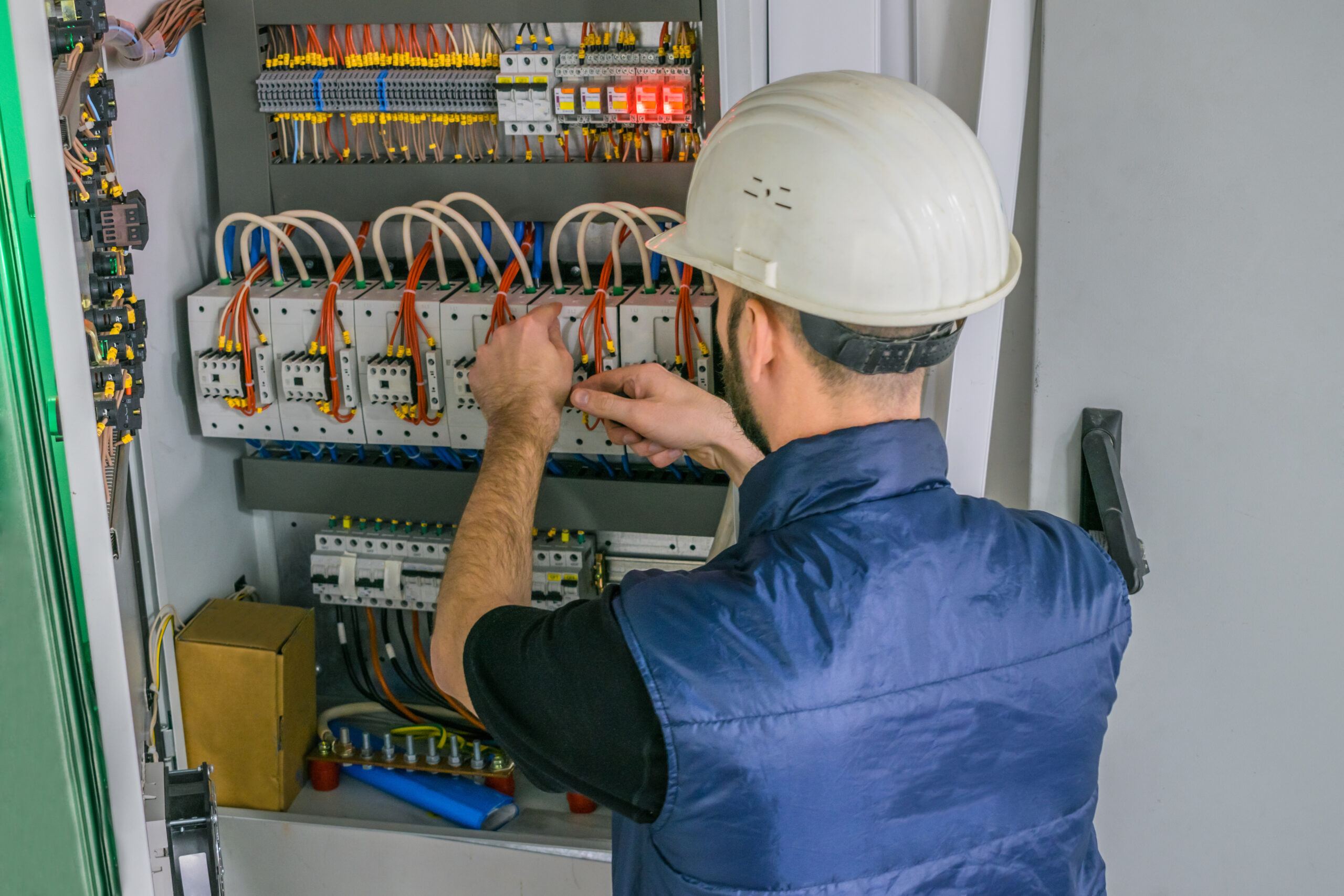 An electrician in a helmet connects the wires of the circuit breakers in the electrical box. A qualified engineer works in an electrical panel. Worker repairs high-voltage equipment.